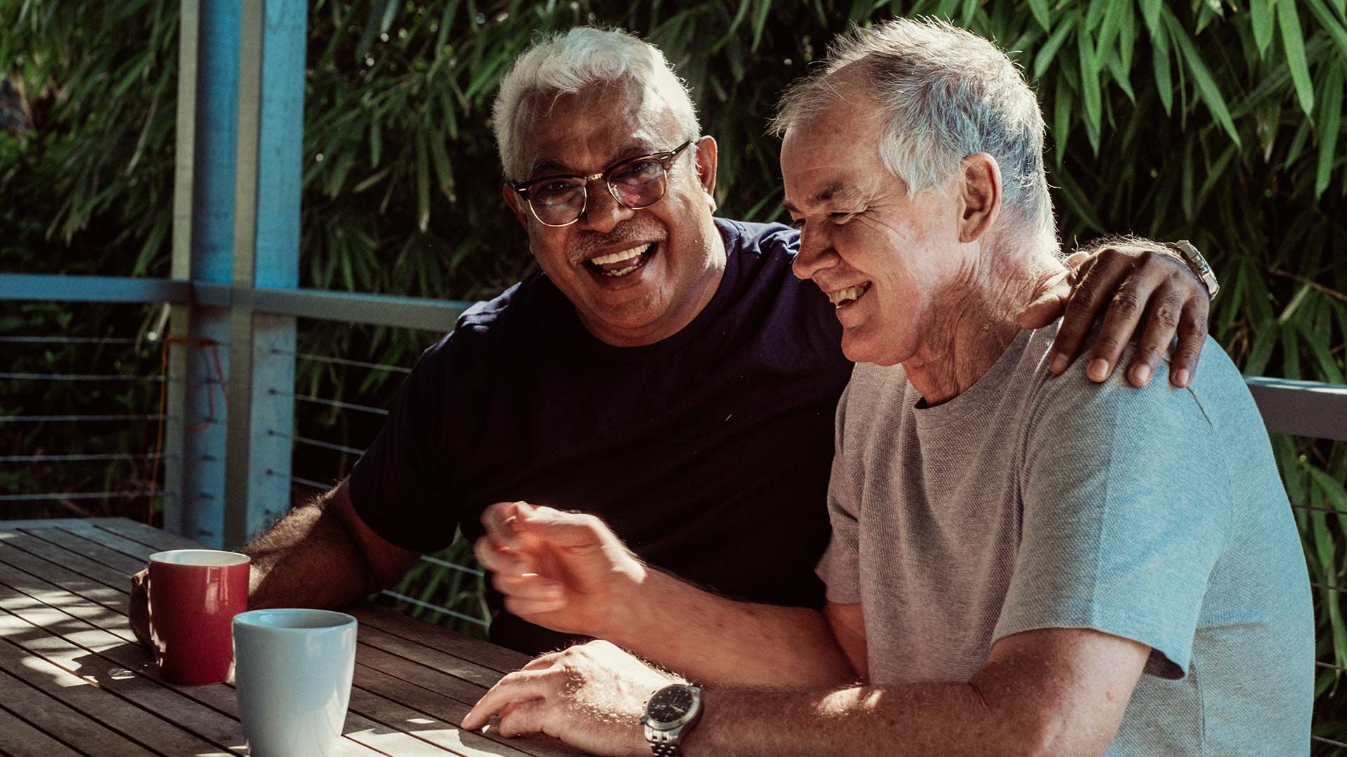 Two men in their 60s sit outside with coffee, both are smiling, and one has his arm around the other