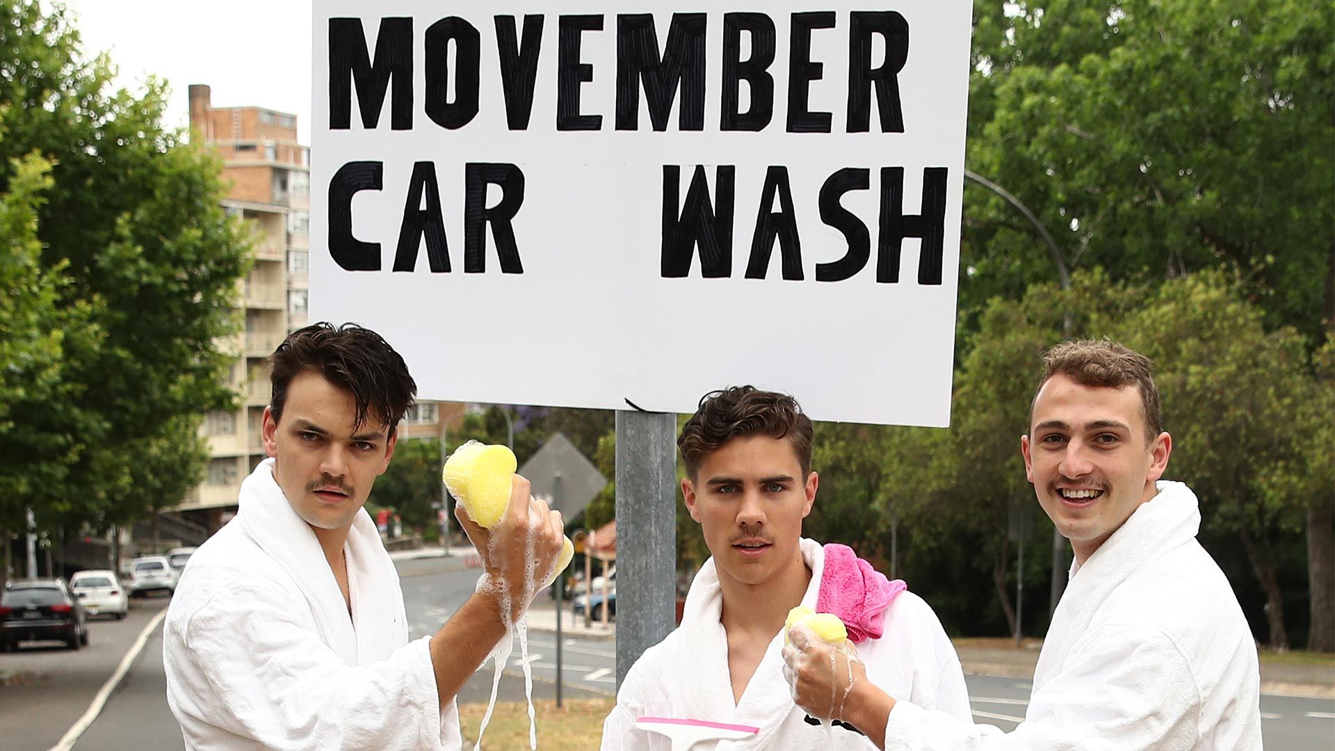 Photo of three men in white dressinggowns, posing on a street. A sign reads: "Movember car wash".